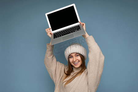 广告浅蓝色摄影照片_照片拍摄的美丽微笑快乐年轻深色金发女性直发在冬天温暖的针织米色帽子拿着电脑笔记本电脑头看着相机穿着米色冬季毛衣隔离在浅蓝色墙壁背景