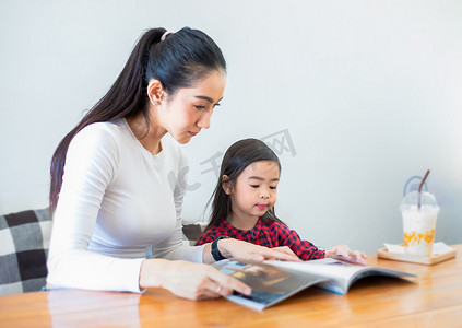 一位亚洲母亲在学期休息期间在客厅的桌子上教女儿读书，家里的桌子上摆着冷牛奶。