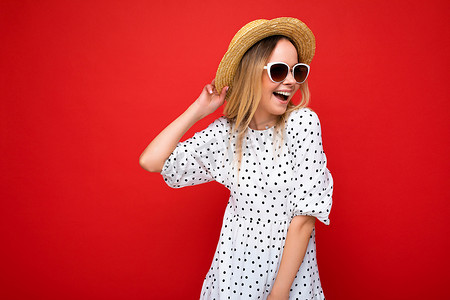 照片中，迷人、微笑的年轻金发女性身穿夏日休闲服，戴着时尚太阳镜，在色彩缤纷的背景墙上望着旁边
