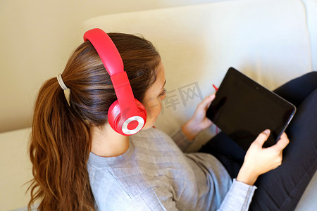 躺在沙发里的女孩摄影照片_快乐的年轻女人在阁楼公寓里戴着耳机躺在沙发上，在平板电脑上选择曲目