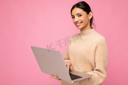 美丽微笑的黑发年轻女子的特写肖像，她拿着上网本电脑，看着粉红色墙壁背景上孤立的相机