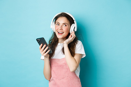 年轻女性戴上耳机并在智能手机上挑选音乐、拿着手机、听歌、站在蓝色背景上的形象