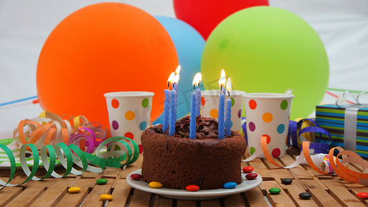 气球墙摄影照片_巧克力生日蛋糕，质朴的木桌上燃烧着蓝色蜡烛，背景是彩色气球、礼物、塑料杯、糖果和背景白墙
