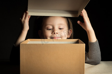 礼物盒子打开摄影照片_情感快乐的微笑小女孩打开一个期待已久的包裹，里面有礼物，复制纸板上的空间，看看盒子