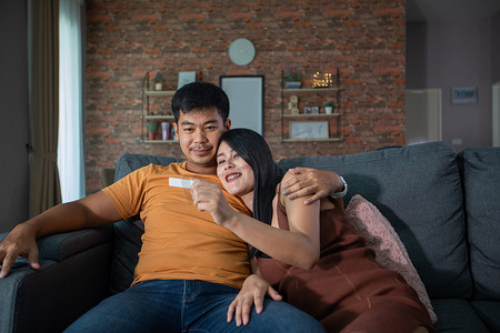 高兴摄影照片_幸福的亚洲夫妇在家里的沙发上进行妊娠测试后，微笑着看起来很高兴
