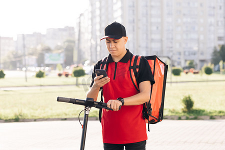 带红色热背包的男性快递送餐员用电动滑板车环顾街道，使用智能手机导航。