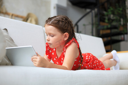 平板游戏摄影照片_微笑的小女孩躺在沙发上玩在线游戏，网上冲浪信息，在平板电脑上使用有趣的应用程序。