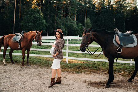 一个怀孕的女孩，在大自然中的一个围场附近的马旁边戴着一顶帽子，肚子很大。穿着棕色连衣裙的时尚孕妇与马。