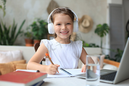 戴着耳机的微笑小女孩在家里使用笔记本电脑在线学习手写，戴着耳机的可爱快乐的小孩在 PC 上上互联网课程或课程。