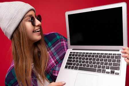 美丽笑笑的金发年轻女子特写肖像拿着电脑笔记本电脑与空显示器屏幕戴着太阳眼镜帽子和彩色衬衫看着上网本显示器和键盘隔离在红墙背景