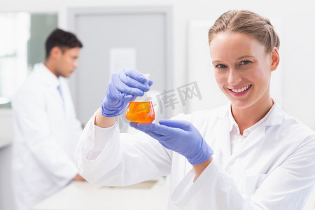橙色实验摄影照片_微笑的科学家看着相机，拿着装有橙色液体的烧杯