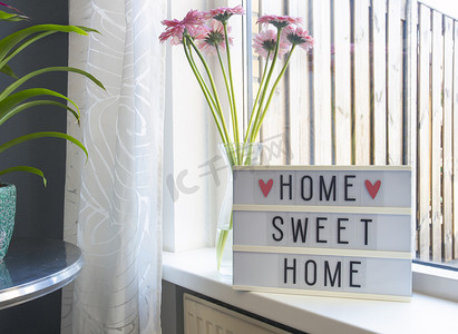 灯箱上的 Sign Home sweet home 文字，靠近窗户的窗台，粉红色的花朵，装饰框架现代室内背景，时尚的房子舒适角落在可爱的客厅