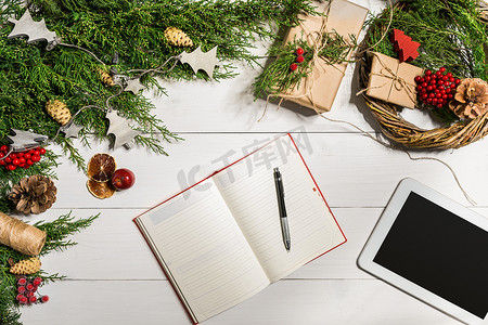 平板电脑和笔记本靠近桌子，配有平板电脑礼品盒、圣诞铃铛和蓬松的冷杉树枝。