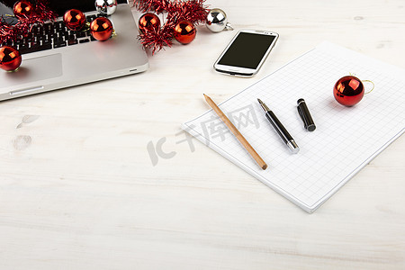 圣诞假期的电脑工作概念：带开放式铝制笔记本电脑的浅色木桌、红色装饰、红色小饰品、智能手机、方块笔记、铅笔和笔在方块笔记上