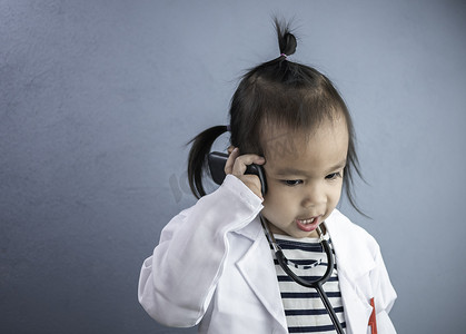 亚洲小女孩角色扮演医生职业，身穿白色长袍制服，面带微笑，通过电话交谈。