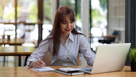 一个穿着白衬衫的年轻开朗的女孩微笑着，微笑着写下笔记，为学生在笔记本电脑桌面桌上进行高管培训
