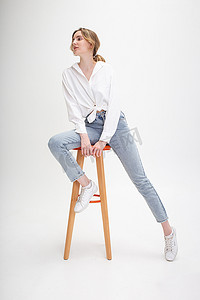 坐在凳子上的人摄影照片_年轻沉思的白人女性穿着衬衫和蓝色牛仔裤，坐在凳子上