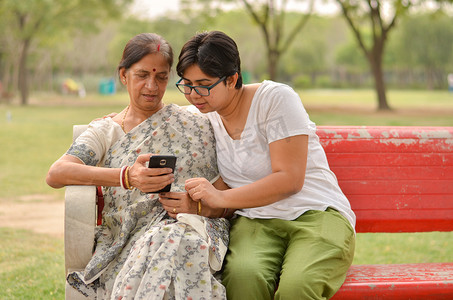 印度新德里的一个公园里，年轻的印度女孩和一位印度孟加拉老妇人坐在红色长凳上看着手机，忙着聊天