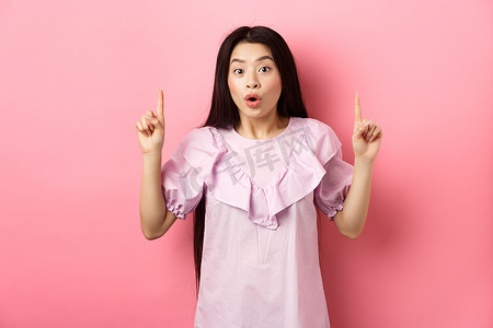 粉色促销背景摄影照片_兴奋的亚洲女孩展示了很棒的促销优惠，指着手指，惊讶地喘着粗气，站在粉色背景上