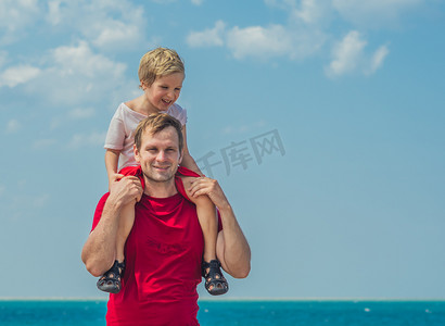 快乐的微笑父亲拥抱在肩膀上男孩男孩站在蓝色拉祖尔海全景天际线地平线阳光天晴朗的天空。