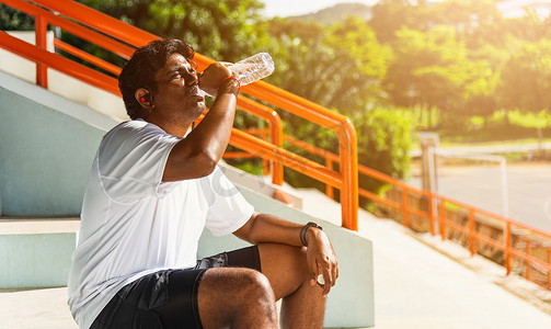 特写亚洲年轻运动跑步者黑人戴着运动员耳机，他在户外街道健康公园跑步后从瓶子里喝水