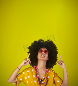 黄色向右指示摄影照片_美丽的女人，留着卷曲的非洲发型，戴着心形太阳镜，用黄色背景上的手指看着并指示向右的方向