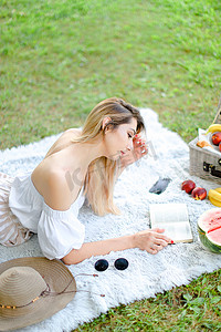 年轻的白人女孩看书，躺在格子上，靠近水果和帽子，背景是草。