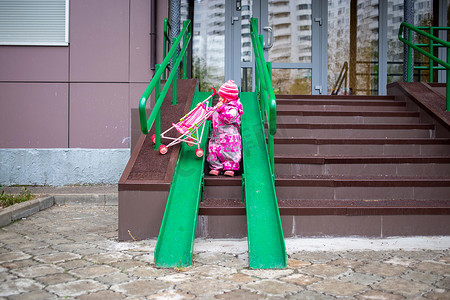 可爱的幼儿带着玩具婴儿车沿着轮椅、手推车和婴儿车的钢栏杆坡道行走。