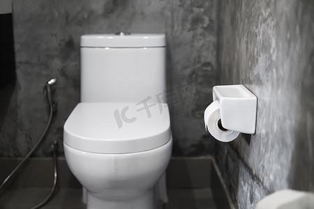家庭浴室白色马桶上的白色悬挂马桶座圈，墙上铺着混凝土风格的灰色瓷砖，墙上挂着卫生纸。