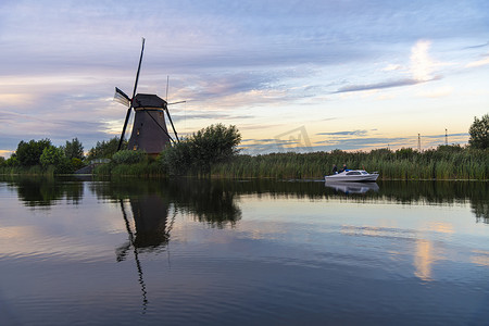 强风吹拂摄影照片_荷兰风车沿着运河铺设，日落时分，强风吹动野草