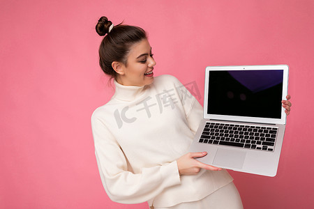 照片中，美丽的黑发女孩穿着白色毛衣，拿着电脑笔记本电脑，看着粉色墙壁背景上隔离的上网本