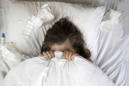 一个生病的年轻女人躺在床上，头盖在毯子下，俯视图旁边放着一堆纸巾。
