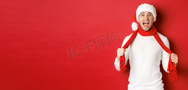 愤怒的成年男子讨厌圣诞节，戴着围巾和圣诞帽，大喊大叫，在红色背景下发疯