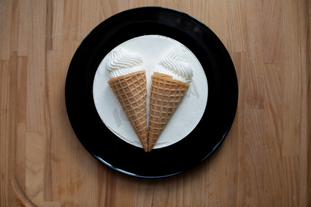 圆形白色甜点甜可口的冰淇淋蛋糕，装饰着两个冰淇淋锥，放在木板背景黑盘上的华夫饼杯里。