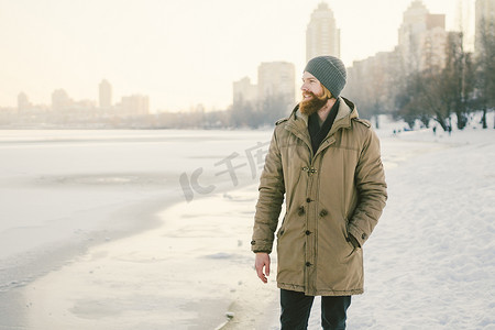 特写白人年轻男性红发和胡须，戴着帽子，穿着公园外套，在湖面背景下摆出冬季模特的姿势，以冻结雪