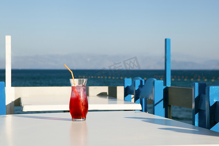 红色果汁摄影照片_杯新鲜的红色果汁在海边的桌子上。