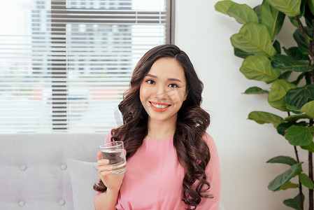 年轻漂亮的亚洲女人在沙发上放松，手里拿着一杯纯净水