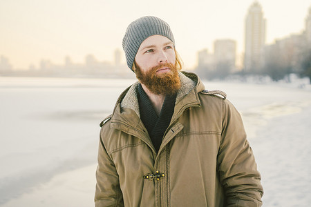 特写白人年轻男性红发和胡须，戴着帽子，穿着公园外套，在湖面背景下摆出冬季模特的姿势，以冻结雪