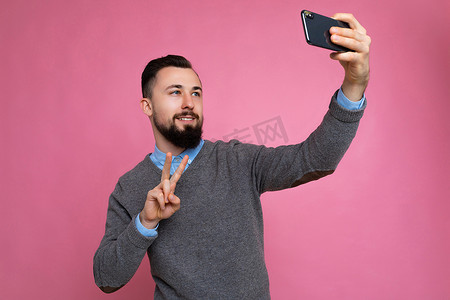 正面英俊的年轻黑发未剃须男子的照片，留着胡子，穿着休闲的灰色毛衣和蓝色衬衫，与粉红色背景墙上隔开，手持智能手机自拍照片，看着手机屏幕显示并展示和平姿态