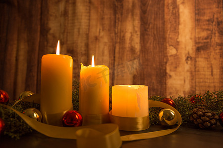 金色的缎带摄影照片_圣诞节概念的温暖：三支蜡烛的特写，在深色木桌上点燃，桌上有松枝、天然松果、金色和红色明亮的小玩意，上面有金色缎带
