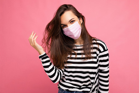 保护抵御摄影照片_年轻漂亮的女人脸上戴着可重复使用的病毒防护粉红色面具，抵御粉红色背景墙上分离出的冠状病毒