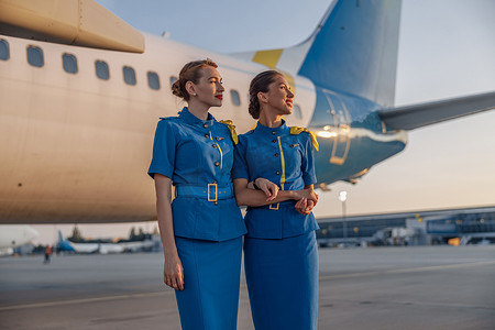 日落时，两位身着蓝色制服的漂亮空姐微笑着站在机场一架大型客机前