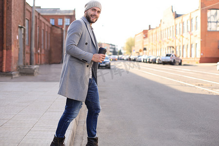 英俊的年轻人穿着灰色外套，戴着帽子，端着一杯咖啡过马路。