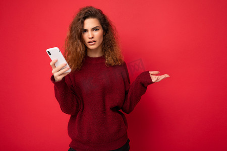 深红色背景摄影照片_照片中，一头卷发、穿着深红色毛衣的漂亮问心烦意乱的年轻女性在红色背景中被隔离，手持手机看着相机