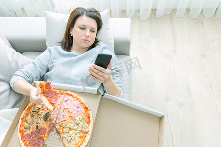 悲伤的女人吃披萨，拿着手机躺在家里的沙发上，蓝色调