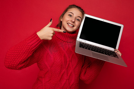 照片中，美丽而满意的快乐年轻女性手持电脑笔记本电脑，看着穿着红色毛衣的相机，显示在红墙背景上的监视器显示屏上