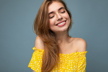 年轻微笑可爱美丽的深色金发女郎的特写照片，她的情感真诚，隔离在背景墙上，有复制空间，穿着时尚的夏季黄色连衣裙。