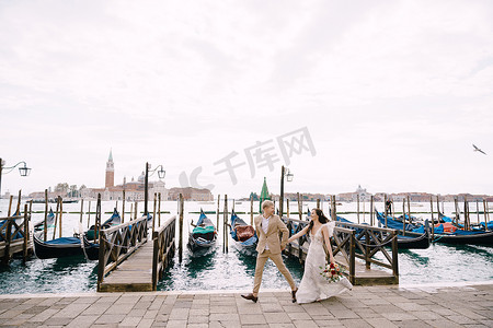 牵手奔跑摄影照片_新娘和新郎沿着贡多拉码头奔跑，在威尼斯牵手，靠近圣马可广场，俯瞰圣乔治马焦雷和落日的天空。