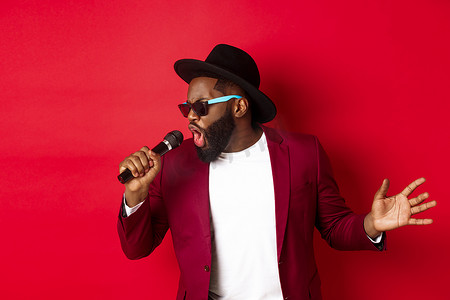 唱歌的男的摄影照片_热情的黑人男歌手在红色背景下表演，对着麦克风唱歌，穿着派对服装，站在红色背景上