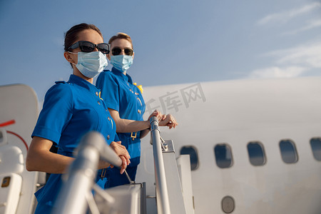白天，两名身着蓝色制服、戴着太阳镜和防护面罩的年轻空姐看着镜头，站在楼梯上